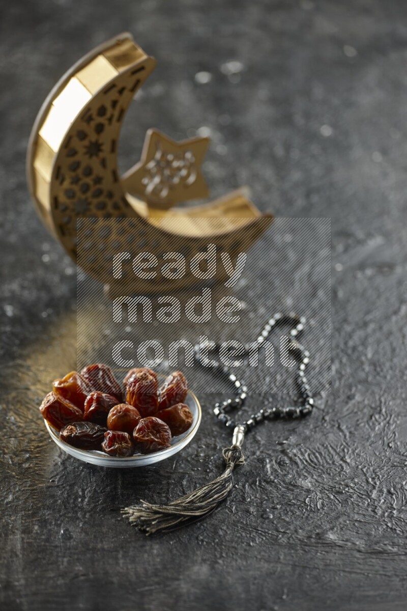 A wooden golden crescent lantern with different drinks, dates, nuts, prayer beads and quran on textured black background