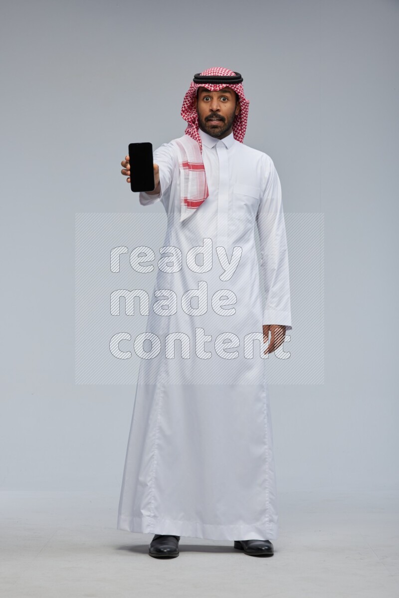 Saudi man Wearing Thob and shomag standing showing phone to camera on Gray background