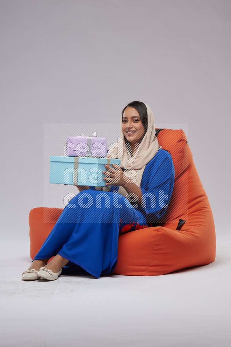 A woman sitting on an orange beanbag wearing Jalabeya holding a gift box
