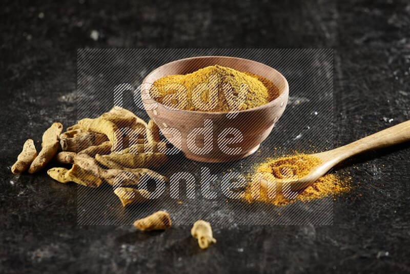 A wooden bowl and a wooden spoon full of turmeric powder with dried turmeric fingers on textured black flooring