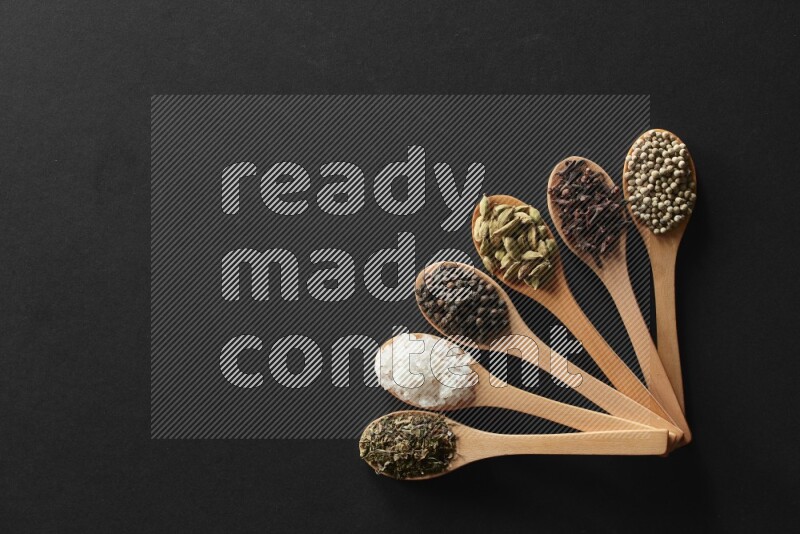 الفلفل الأبيض والقرنفل والهيل والملح والفلفل الأسود وريحان مجفف في معالق خشبية علي خلفية سوداء