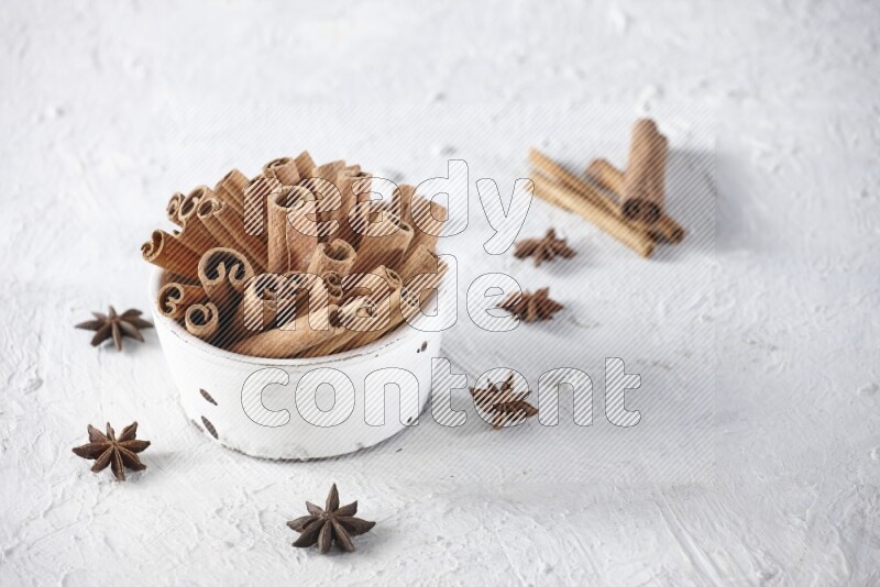 White bowl full of cinnamon sticks surrounded by star anis on a textured white background in different angles