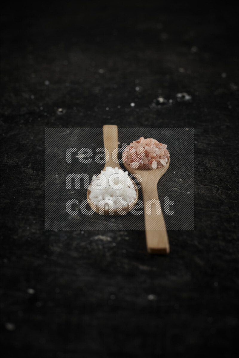 2 wooden spoons filled with fine and coarse salt on black background
