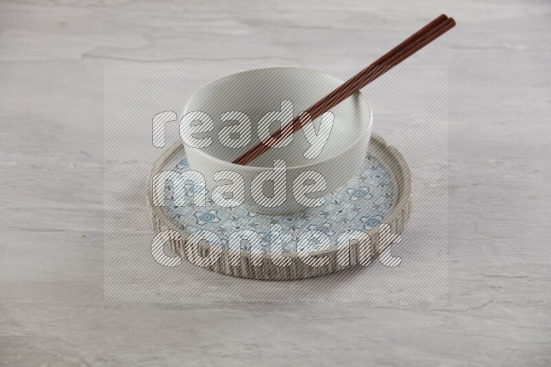 white pottery round bowl on top of multi color round ceramic plate and chopsticks, on grey textured countertop