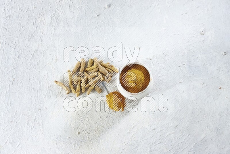 A beige pottery bowl and metal spoon full of turmeric powder and dried turmeric fingers next of them on textured white flooring