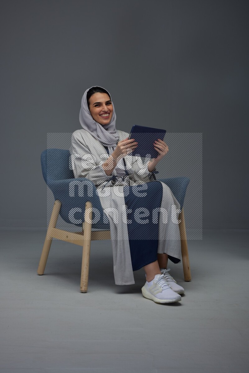 A Saudi female wearing a light gray Abaya and head scarf sitting on a dark grey chair while holding her iPad on a grey background