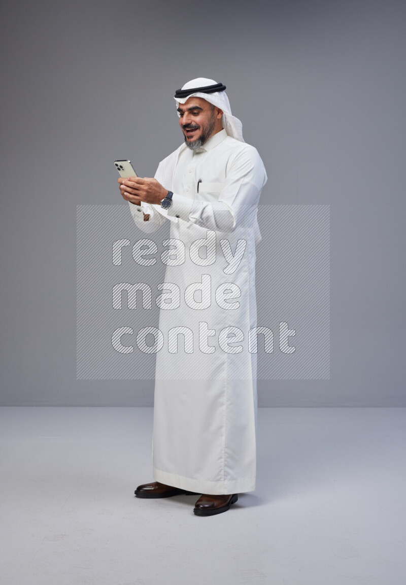 Saudi man Wearing Thob and white Shomag standing texting on phone on Gray background