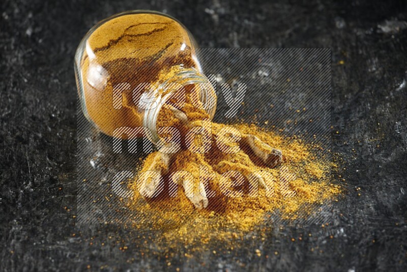 A flipped glass spice jar full of turmeric powder and powder spilled out of it with dried whole fingers on textured black flooring