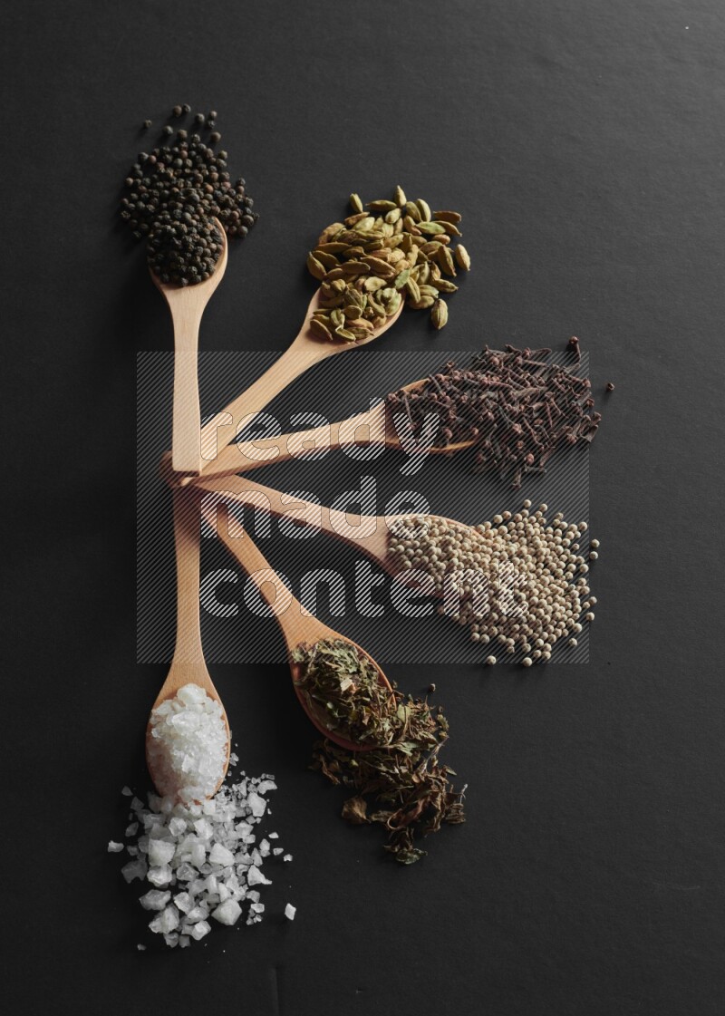 wooden spoons filled with white peppers, cloves, cardamom, salt, black peppers and basil on black flooring