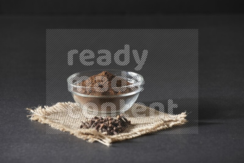 A glass bowl full of cloves powder with cloves grains on a burlap piece on a black flooring