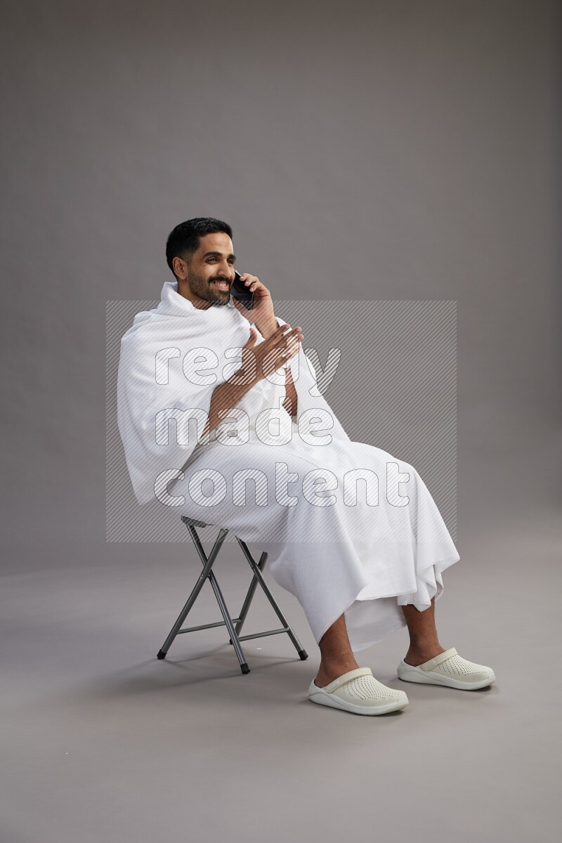 A man wearing Ehram sitting on chair talking on the phone on gray background