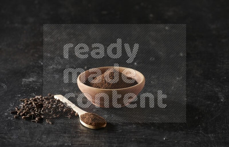 A wooden bowl and a wooden spoon full of cloves powder with spreaded cloves on a textured black flooring