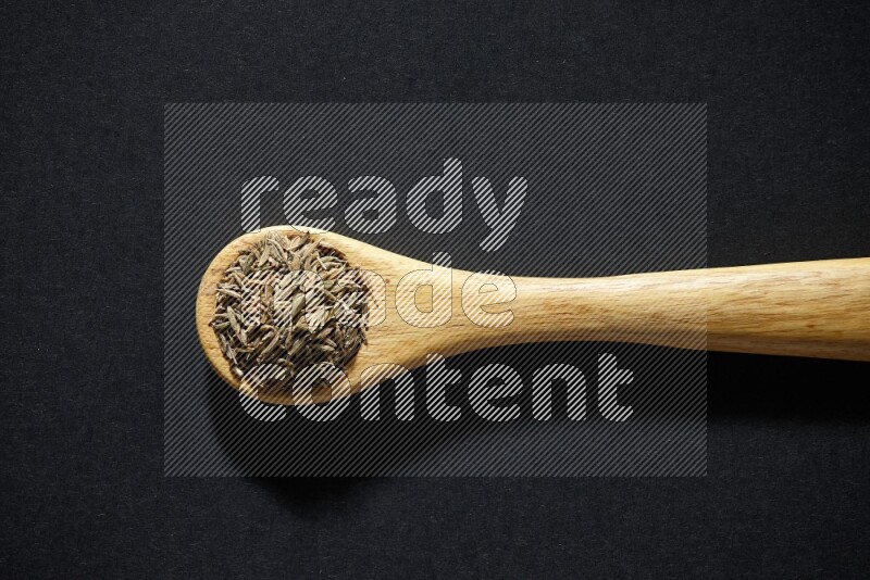 A wooden spoon full of cumin seeds on black flooring