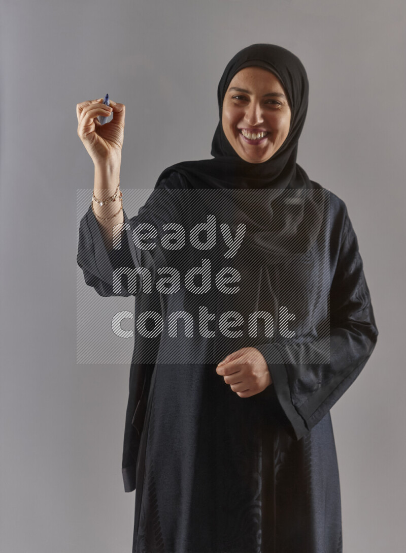 Woman in black abaya and hijab holding a marker pen to write on a transparent board with different reactions and expressions on grey background