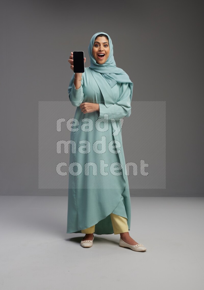 Saudi Woman wearing Abaya standing showing phone to camera on Gray background
