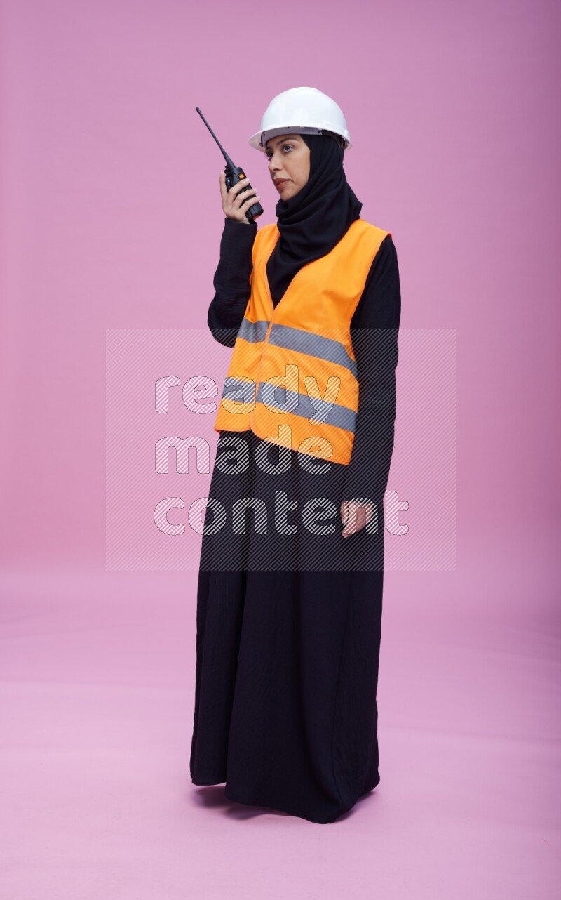 Saudi woman wearing Abaya with engineer vest and helmet standing holding walkie-talkie on pink background