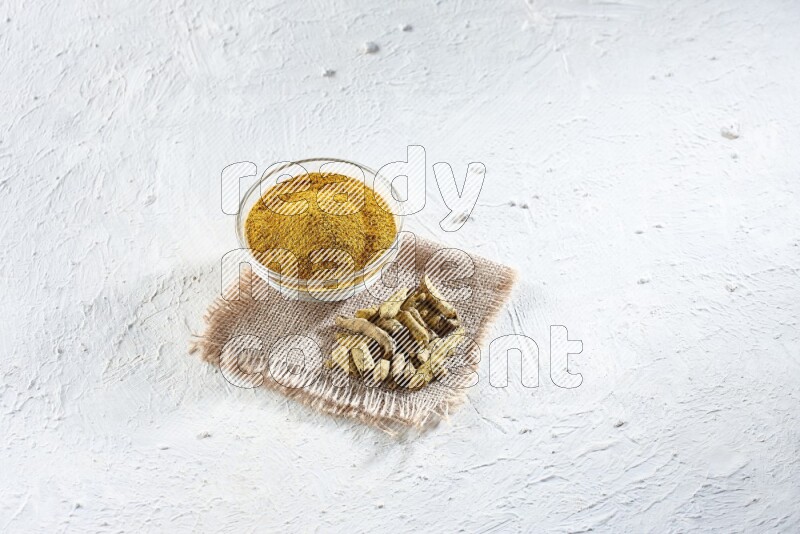 A glass bowl full of turmeric powder and dried turmeric whole finger on a piece of burlap on a textured white flooring
