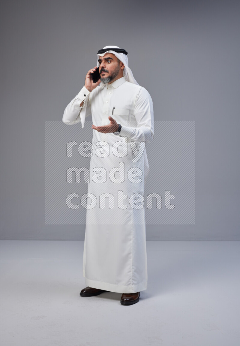 Saudi man Wearing Thob and white Shomag standing talking on phone on Gray background