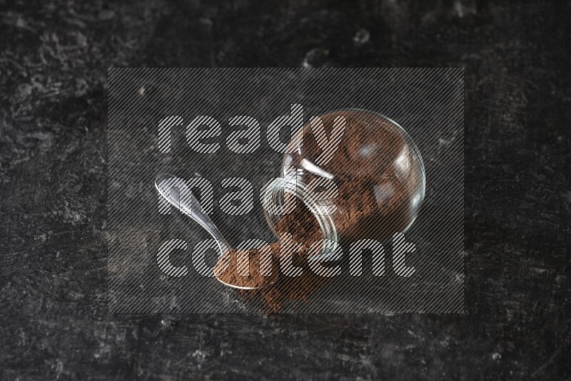 A flipped glass spice jar and a metal spoon full of cloves powder on textured black flooring