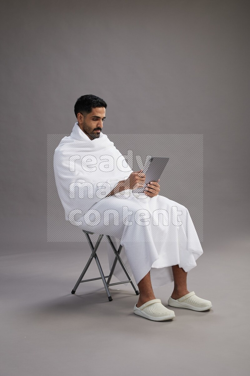 A man wearing Ehram sitting on chair holding a tablet on gray background