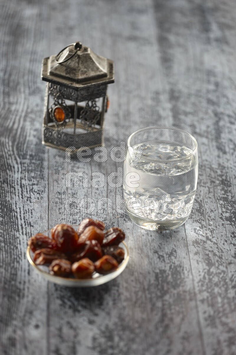 A silver lantern with different drinks, dates, nuts, prayer beads and quran on grey wooden background