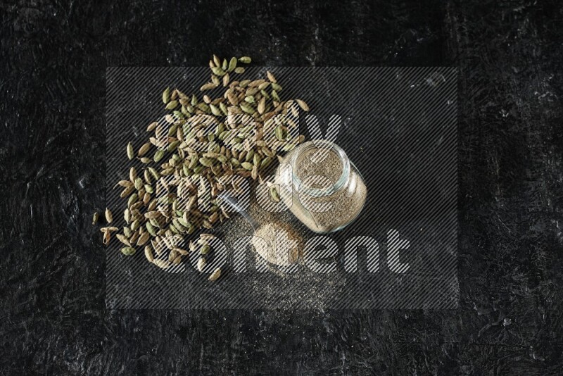 A glass spice jar and a metal spoon full of cardamom powder and cardamom seeds spreaded on textured black flooring