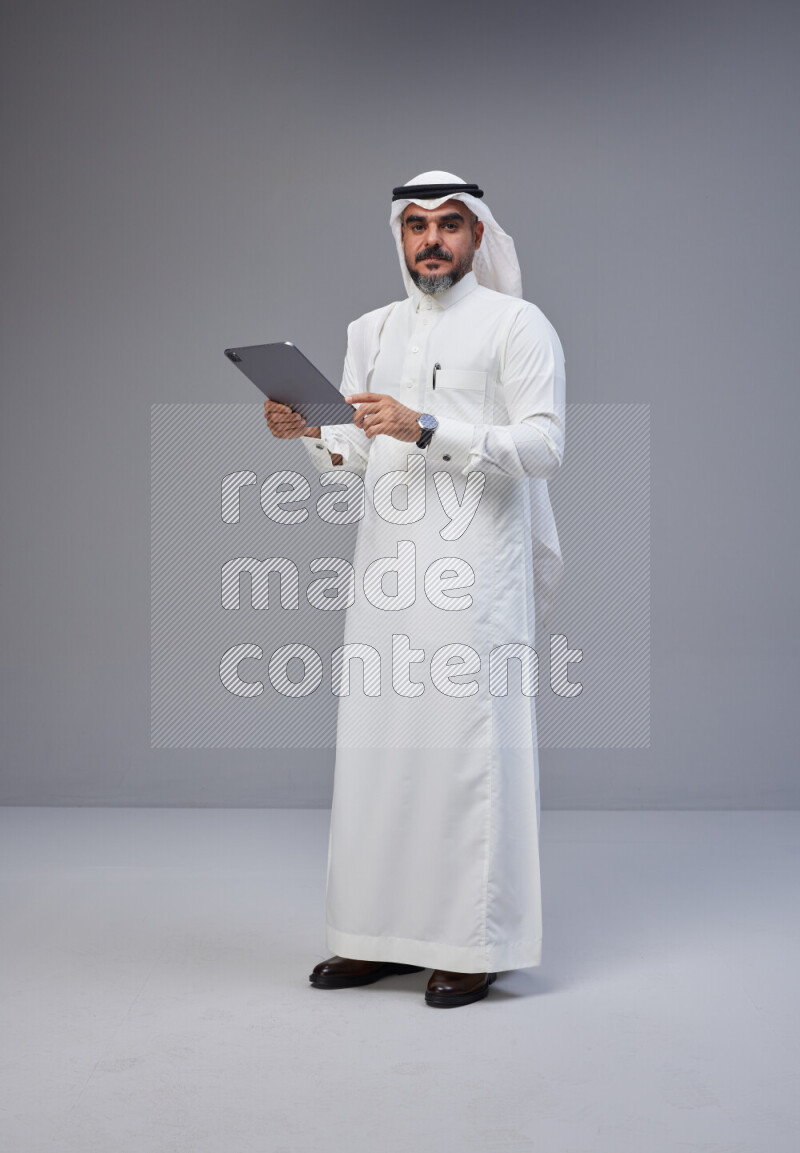 Saudi man Wearing Thob and white Shomag standing working on tablet on Gray background