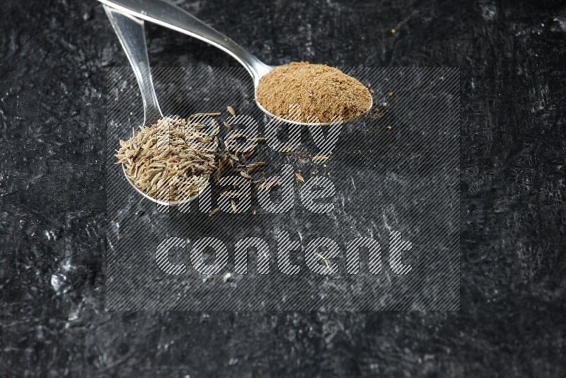 2 metal spoons full of cumin seeds and cumin powder on a textured black flooring