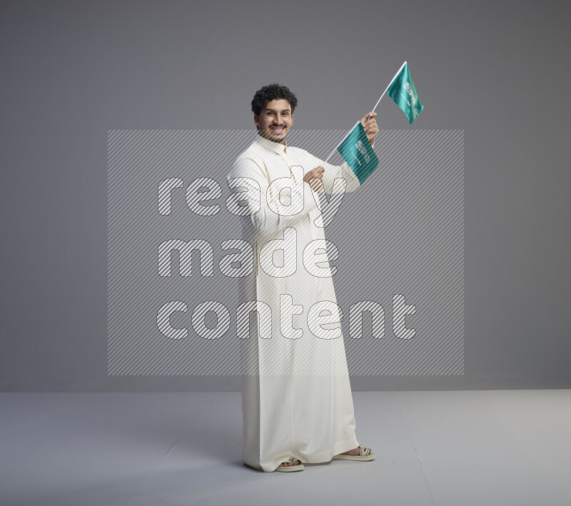 A Saudi man standing wearing thob holding small Saudi flag on gray background
