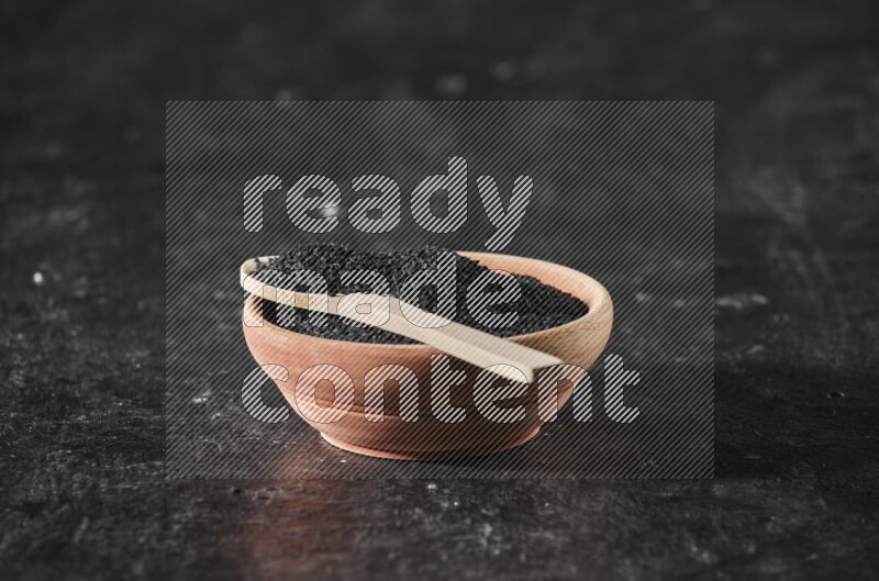 A wooden bowl full of black seeds with wooden spoon full of the seeds on it on a textured black flooring