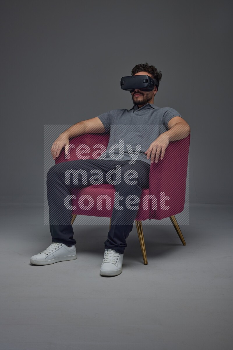 A man wearing casual and wearing VR while sitting on a burgundy chair eye level on a gray background