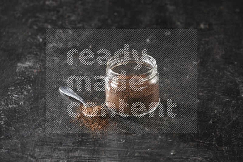 A glass jar full of cloves powder with a metal spoon on a textured black flooring