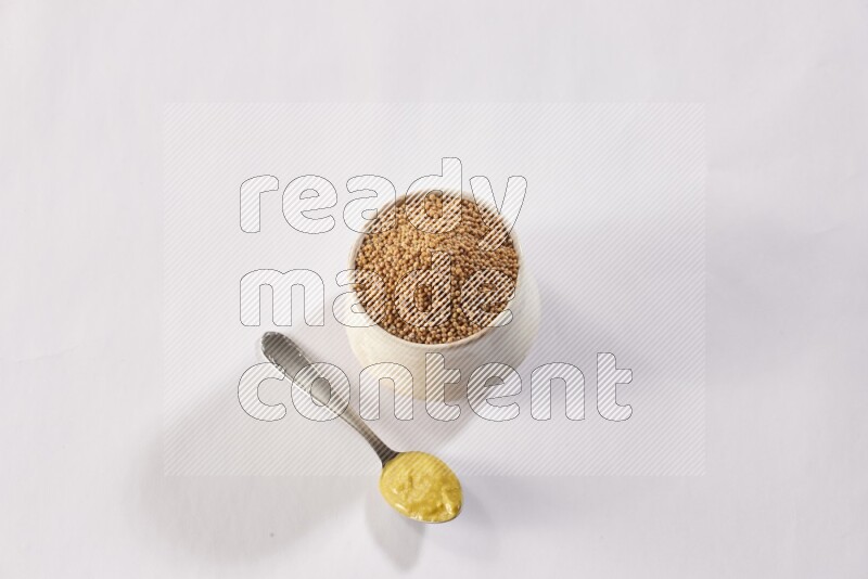A beige pottery bowl full of mustard seeds and a metal spoon full of mustard paste on white flooring