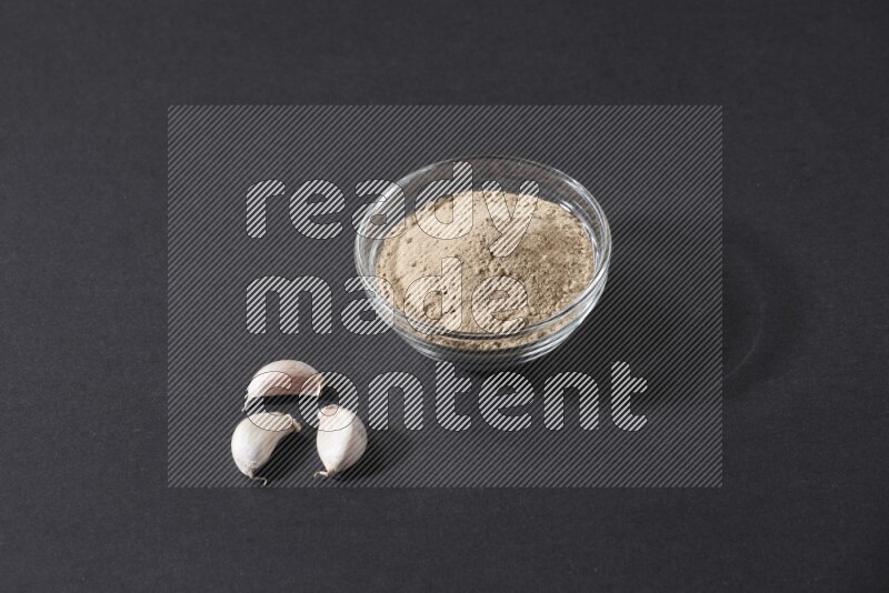 A glass bowl full of garlic powder with garlic bulb and some cloves beside it on a black flooring