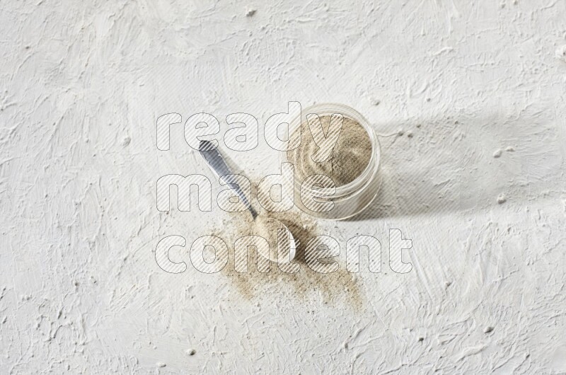 A glass jar and a metal spoon full of white pepper powder on textured white flooring