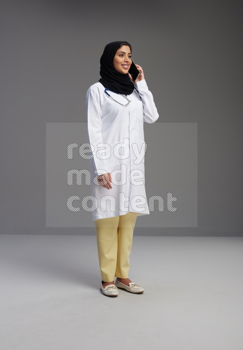 Saudi woman wearing lab coat with stethoscope standing talking on phone on Gray background