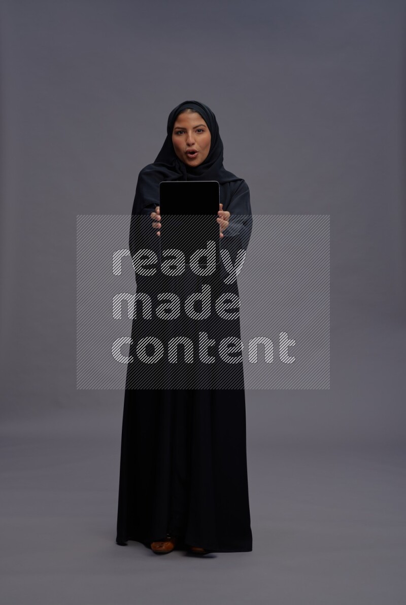 Saudi woman wearing Abaya standing showing tablet to camera on gray background