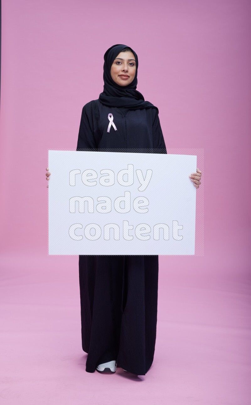 Saudi woman wearing pink ribbon on Abaya standing holding board on pink background