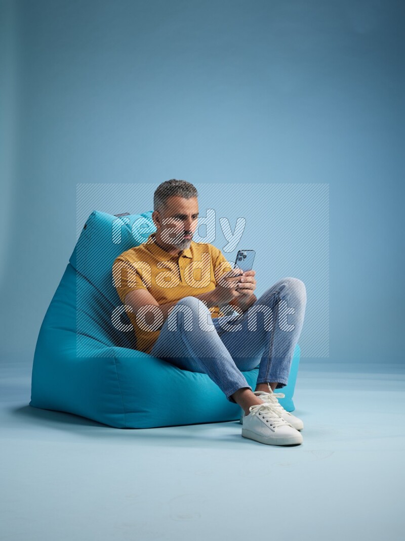 A man sitting on a blue beanbag and texting on phone