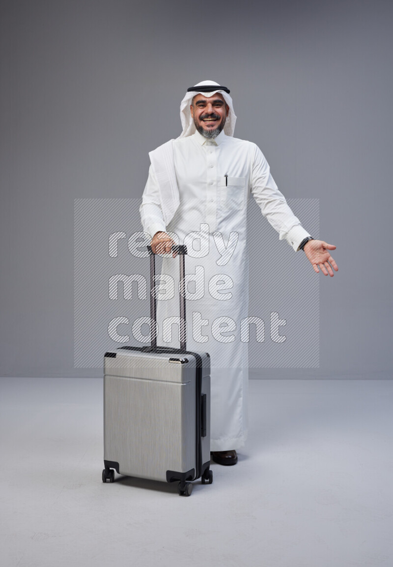 Saudi man wearing Thob and white Shomag standing holding Travel bag on Gray background