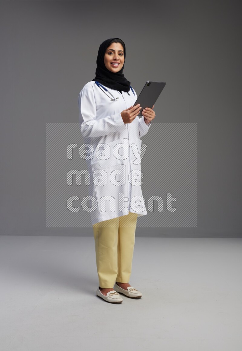 Saudi woman wearing lab coat with stethoscope standing working on tablet on Gray background