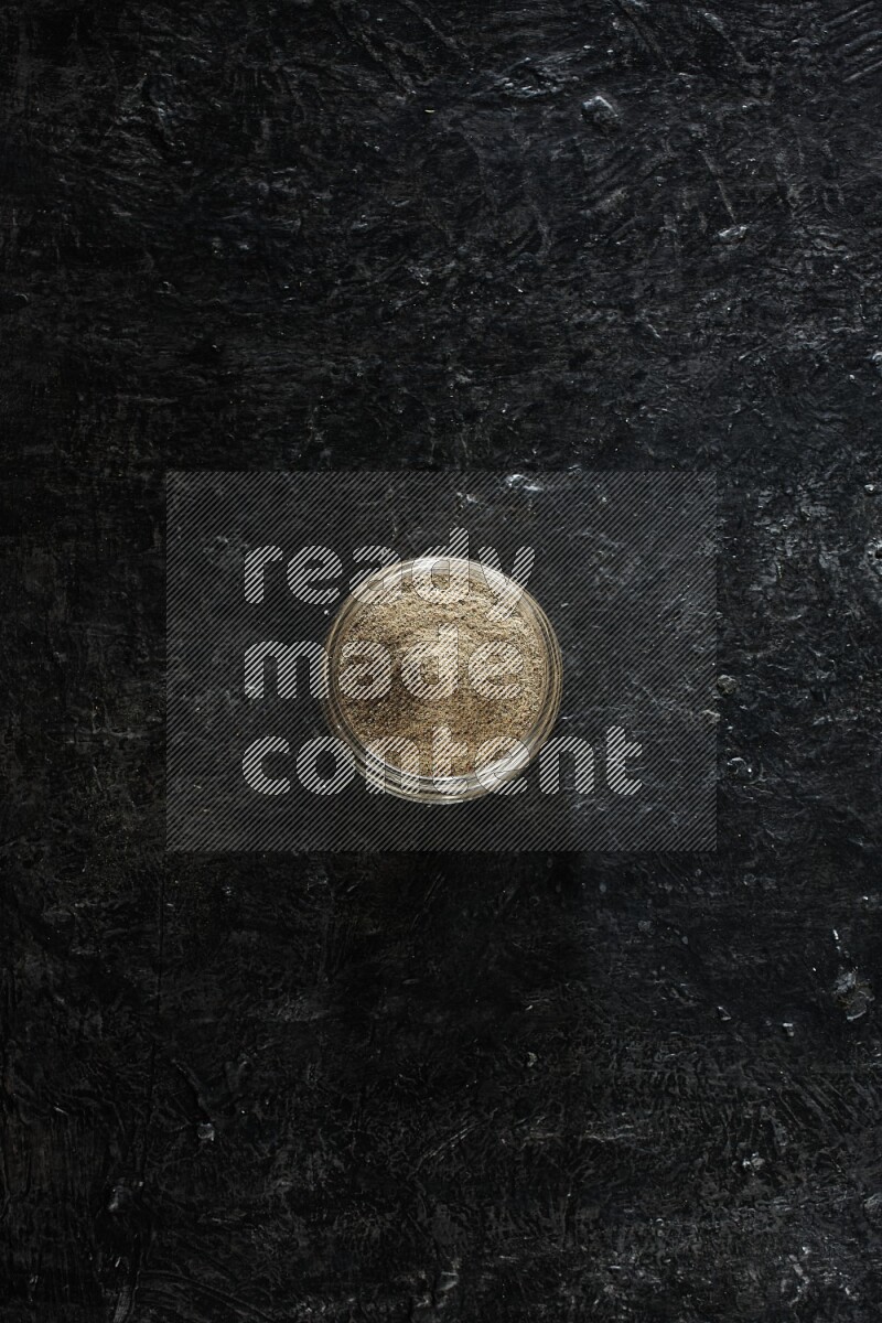 A glass jar full of cardamom powder on textured black flooring
