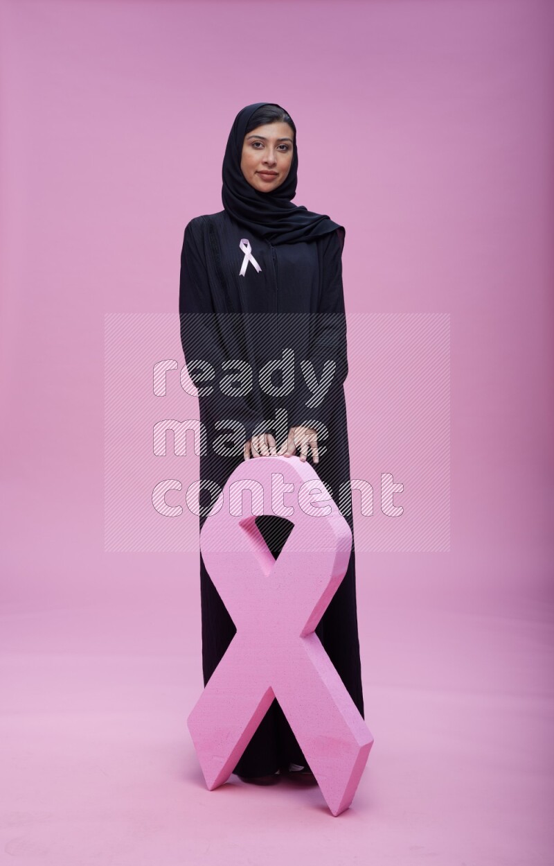 Saudi woman wearing pink ribbon on abaya standing holding awareness ribbon on plain pink background
