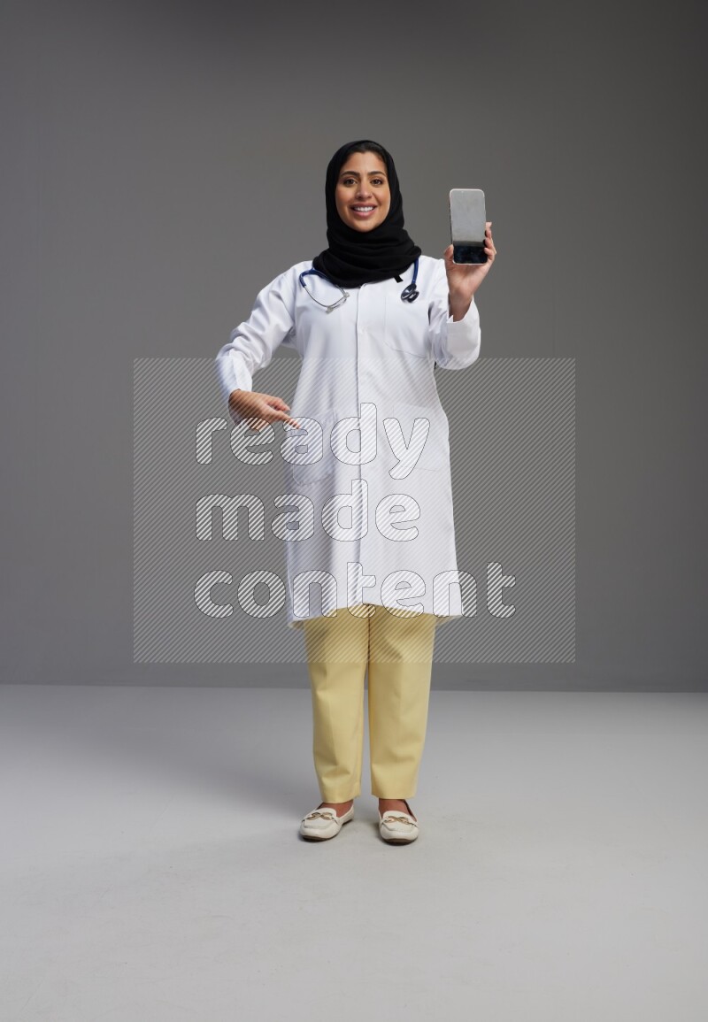 Saudi woman wearing lab coat with stethoscope standing showing phone to camera with sign in the back on Gray background