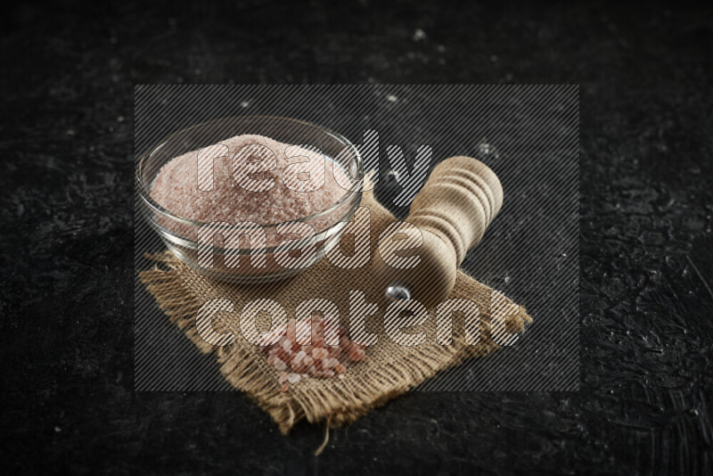 A glass bowl full of pink himalayan salt with a wooden grinder on a burlap fabric all on black background