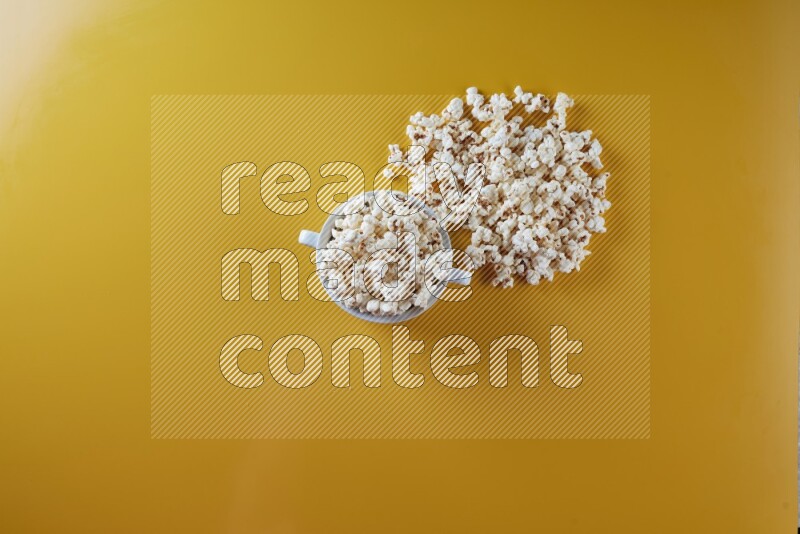 A white ceramic bowl full of popcorn with popcorn beside it on a yellow background in a top view shot