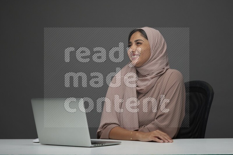 A Saudi woman Setting on her desk on a Gray Background wearing Brown Abaya with Hijab