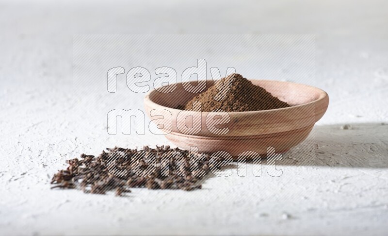 A wooden bowl full of cloves powder with whole cloves beside it on a textured white flooring