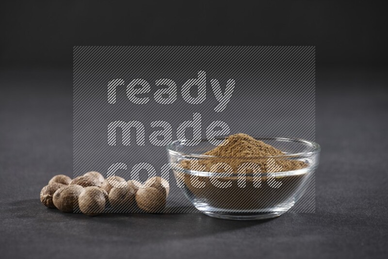 A glass bowl full nutmeg powder with whole seeds beside it on a black flooring