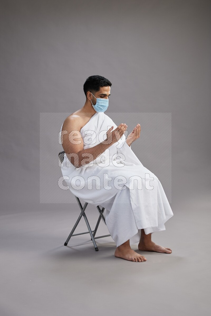 A man wearing Ehram with face mask sitting on chair dua'a on gray background